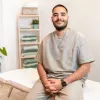 A massage therapist sits on the edge of a massage table, smiling for a photo.