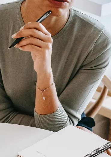 A woman in a long-sleeve shirt holds a black pen to her mouth as she thinks.